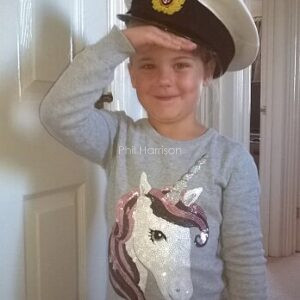 Young girl smiling and saluting, wearing captain's hat and sequinned unicorn jumper.